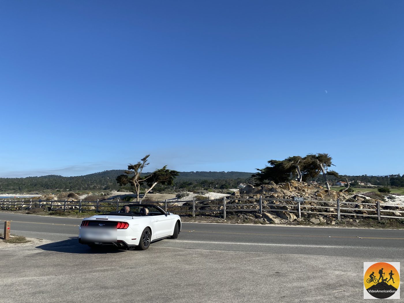 A white convertible pulled over beside windswept cypress trees along 17 Mile Drive, with the moon visible in a blue sky